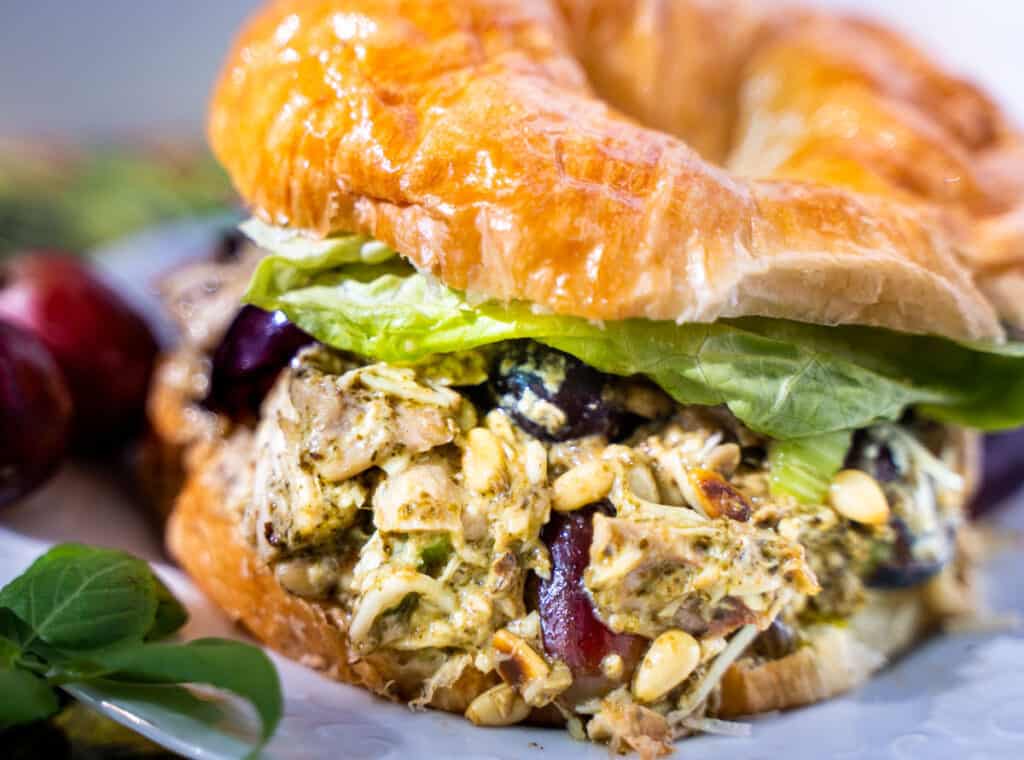 picnic perfect pesto chicken salad on a croissant sitting on a white plate garnished with red grapes and basil