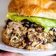 A hand holding the Picnic Perfect Pesto Chicken Salad croissant sandwich on a white plate