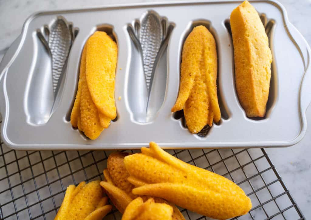 pile of corn eared shaped corn bread on a cooling rack with the baking pan resting next to the corneared shaped cornbread