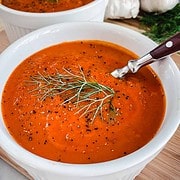 2 white bowls filled with Tomato and Fennel Soup garnished with pepper and a fennel sprig sitting on a wood table with garlic bulbs in the background