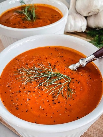 2 white bowls filled with Tomato and Fennel Soup garnished with pepper and a fennel sprig sitting on a wood table with garlic bulbs in the background