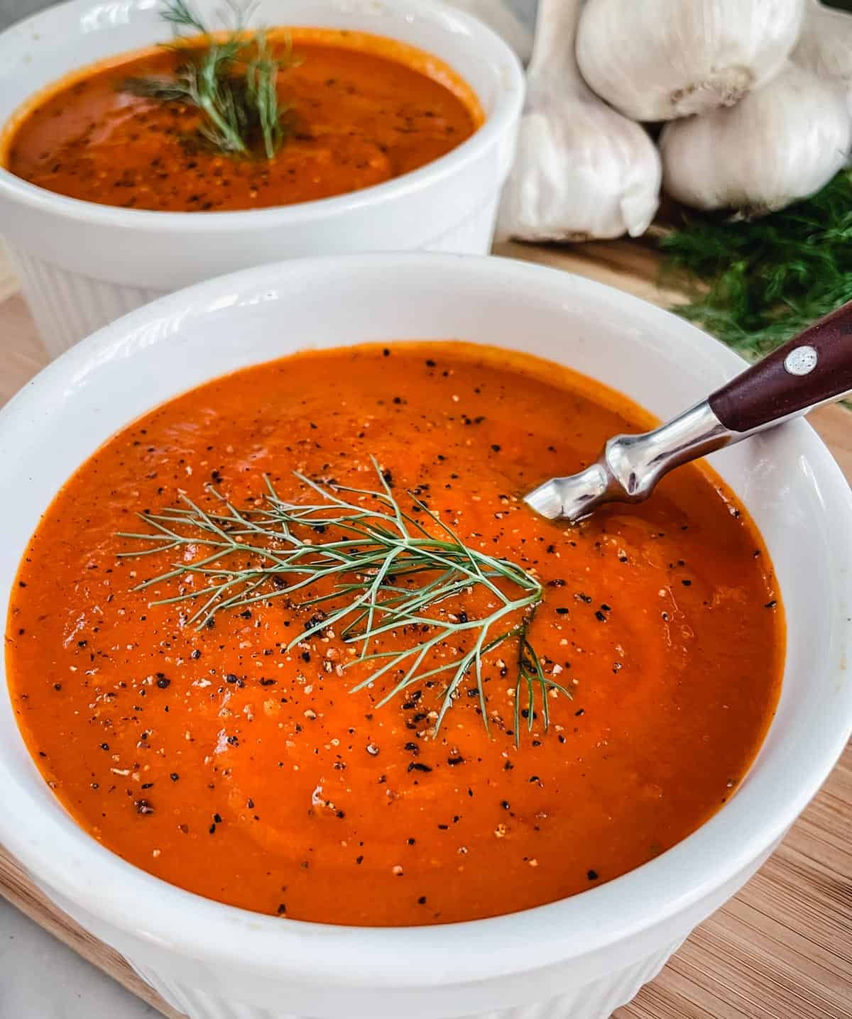 2 white bowls filled with Tomato and Fennel Soup garnished with pepper and a fennel sprig sitting on a wood table with garlic bulbs in the background