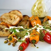 Burrata appetizer with grilled mini orange and red peppers, that has been drizzled with a green basil dressing arranged on a white platter alongside grilled bread cut on the bias and a salad dressing beaker filled with vinaigrette