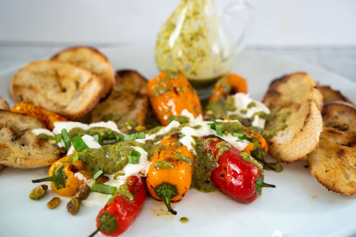 Burrata appetizer with grilled mini orange and red peppers, that has been drizzled with a green basil dressing arranged on a white platter alongside grilled bread cut on the bias and a salad dressing beaker filled with vinaigrette
