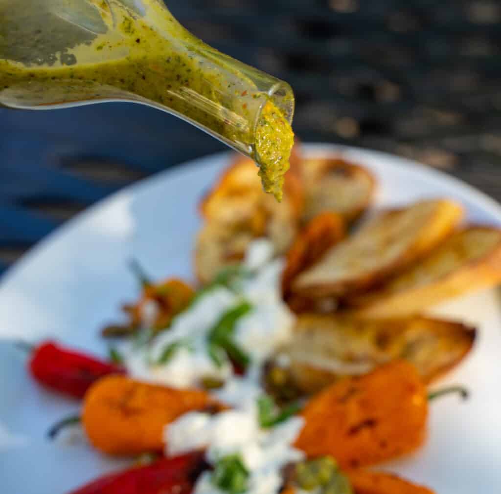 Green basil pistachio vinaigrette in a clear beaker is being drizzled over red and orange peppers, burrato that has been arranged around the peppers.