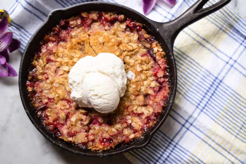 Baked Rhubarb and Cherry Crisp with golden brown oat crisp topping in a cast iron skillet topped with a scoop of vanilla ice cream sitting on a blue and yellow striped dish towel