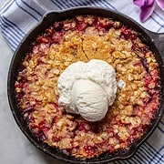 Baked Rhubarb and Cherry Crisp with golden brown oat crisp topping in a cast iron skillet topped with a scoop of vanilla ice cream sitting on a blue and yellow striped dish towel