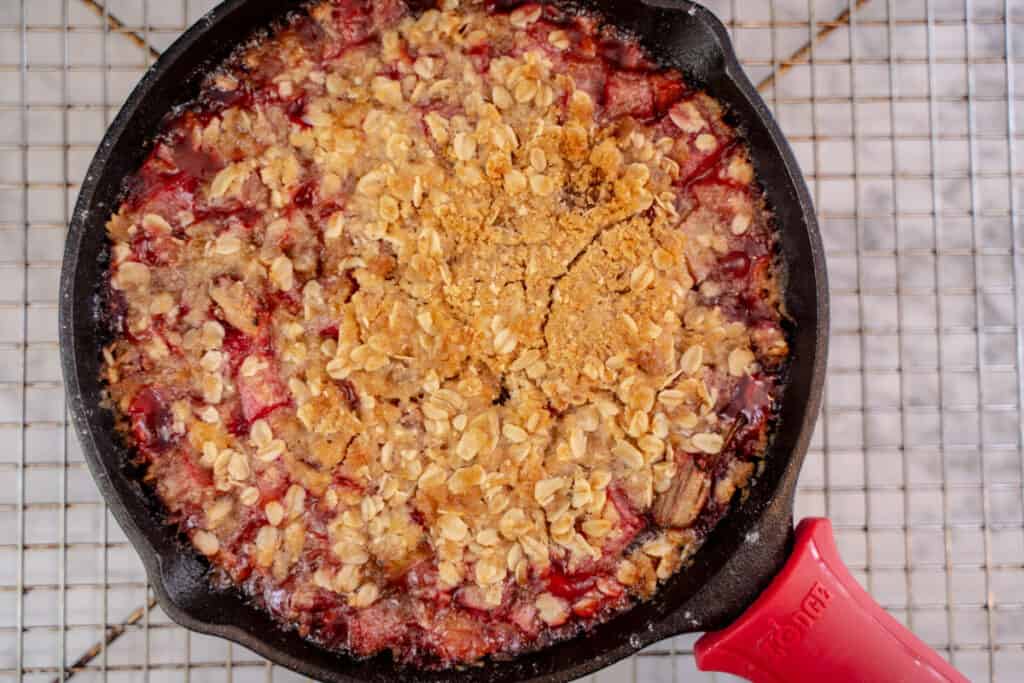 Baked Rhubarb and Cherry Crisp with golden brown oat crisp topping in a cast iron skillet right out of the oven sitting on a baking rack to cool