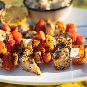 Grilled Swordfish, orange bell pepper and cherry tomatoes on metal skewers sitting on a white platter along side a charred lemon half, cilantro-lime rice in a small bowl, and black beans with pepper and cilantro garnish in a small bowl