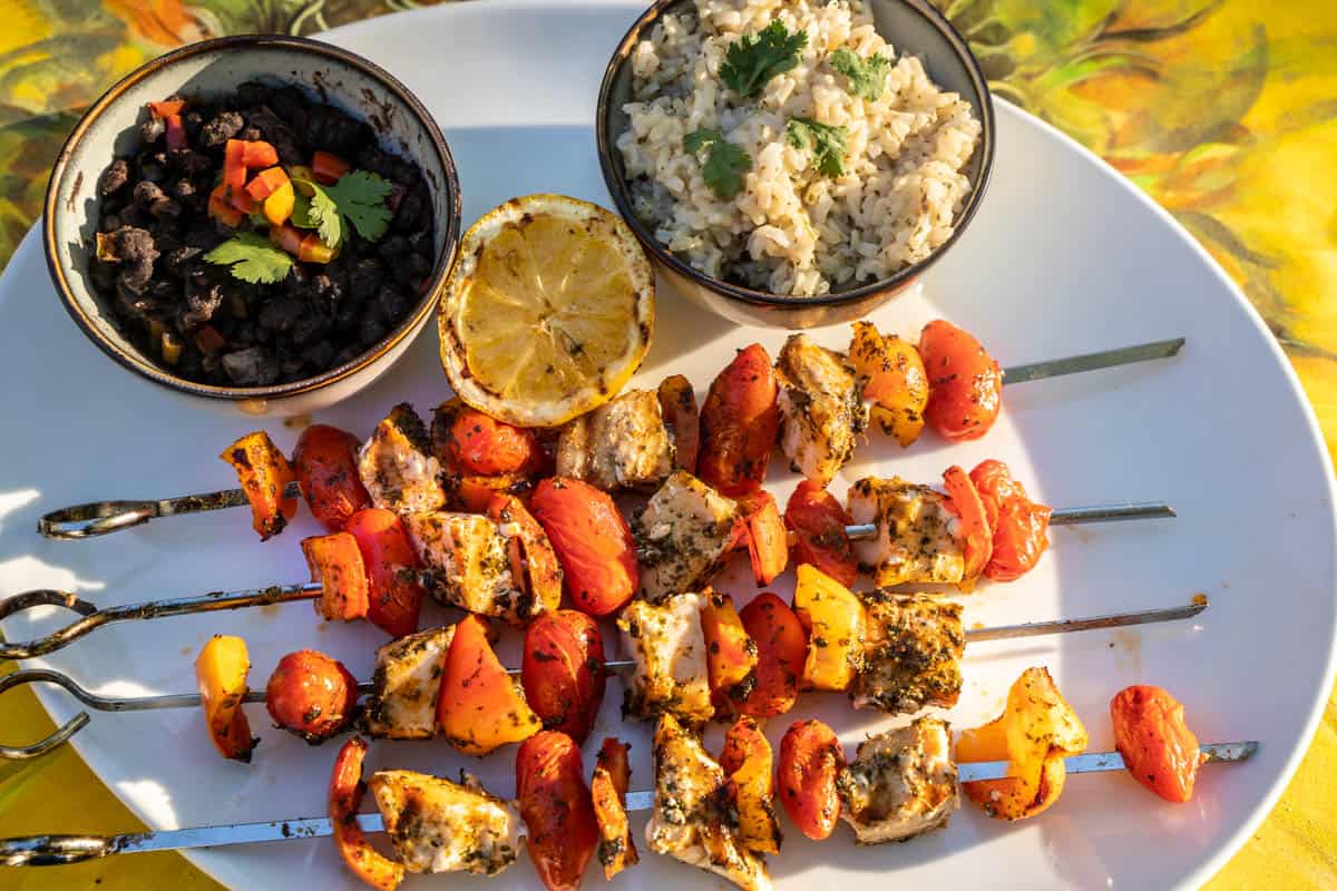 Grilled Swordfish, orange bell pepper and cherry tomatoes on metal skewers sitting on a white platter along side a charred lemon half, cilantro-lime rice in a small bowl, and black beans with pepper and cilantro garnish in a small bowl