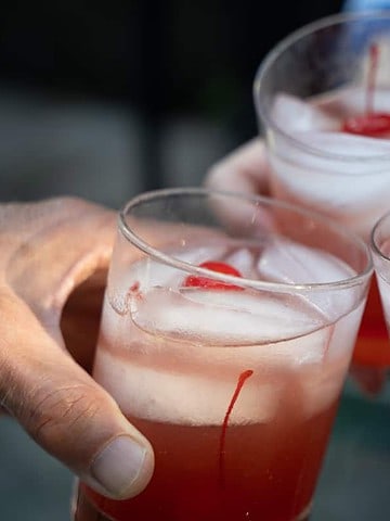 whiskey sour recipe poured over ice in 3 old fashioned glasses and topped with maraschino cherries being held by 3 people for a toast