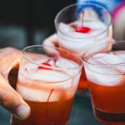 Whiskey Sour Drinks in three cocktail glass over ice garnished with a maraschino cherry held by three hands for a toast.
