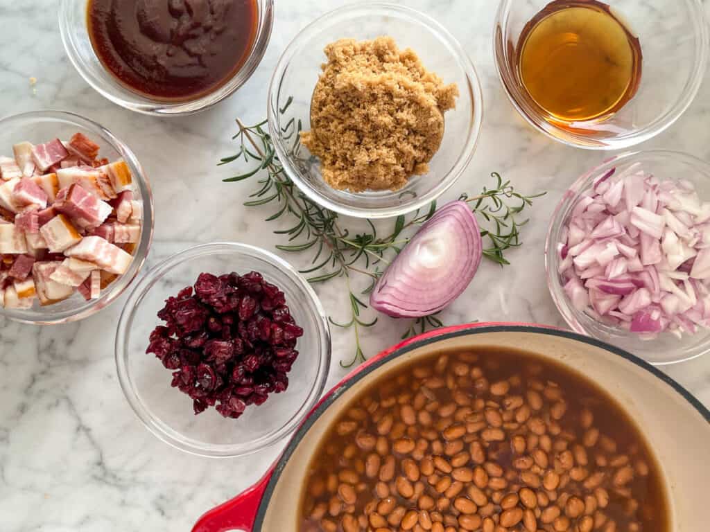 BBQ Baked beans recipe ingredients in individual bowls - baked beans, red onion, brown sugar, maple syrup, cranberries, BBQ sauce, diced bacon