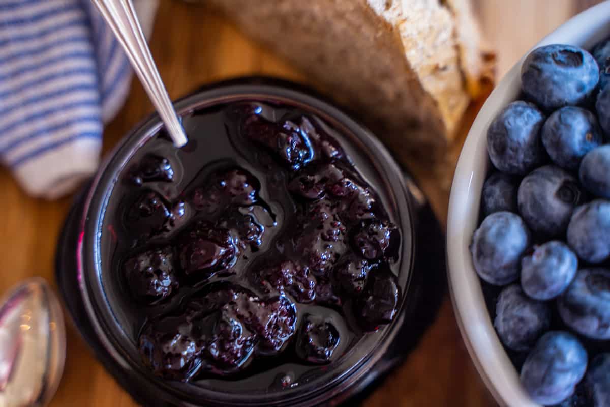 Blueberry jam in a mason jar, fresh blueberries in a white bowl and sourdough sitting on a table