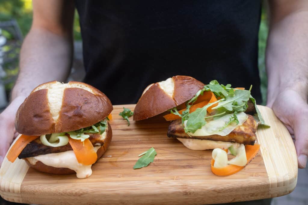 2 Crispy Halloumi Burgers garnished with ribbons of carrots and cucumber, slathered in a sweet chili mayo sauce on a cutting board held by a person