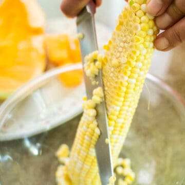 removing kernels with a knife from a ear of corn sitting in a bowl.