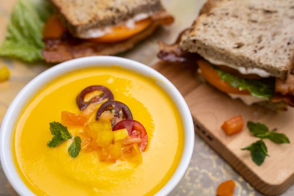 Cantaloupe and Sweetcorn gazpacho in a bowl alongside a BLT sandwich on a cutting boardn