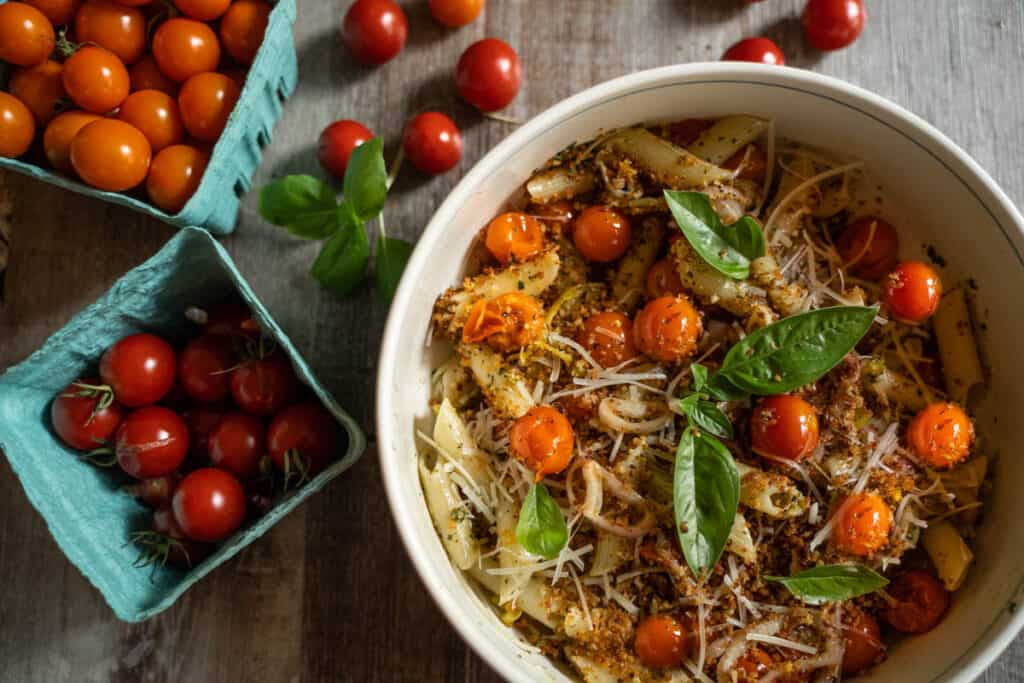 Roasted Cherry Tomato Pasta Recipe for two tossed with basil pesto, roasted garlic, roasted shallots and fried pepperoncini panko topping in a white pasta bowl surrounded by pints of cherry tomatoes