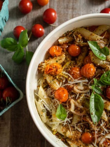 Roasted Cherry Tomato Pasta Recipe for two tossed with basil pesto, roasted garlic, roasted shallots and fried pepperoncini panko topping in a white pasta bowl surrounded by pints of cherry tomatoes