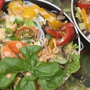 spring roll ingredients of shaved carrots, slices of yellow and red pepper, mushrooms, peanuts, green onions, rice noodles in a bowl drizzled with peanut sauce