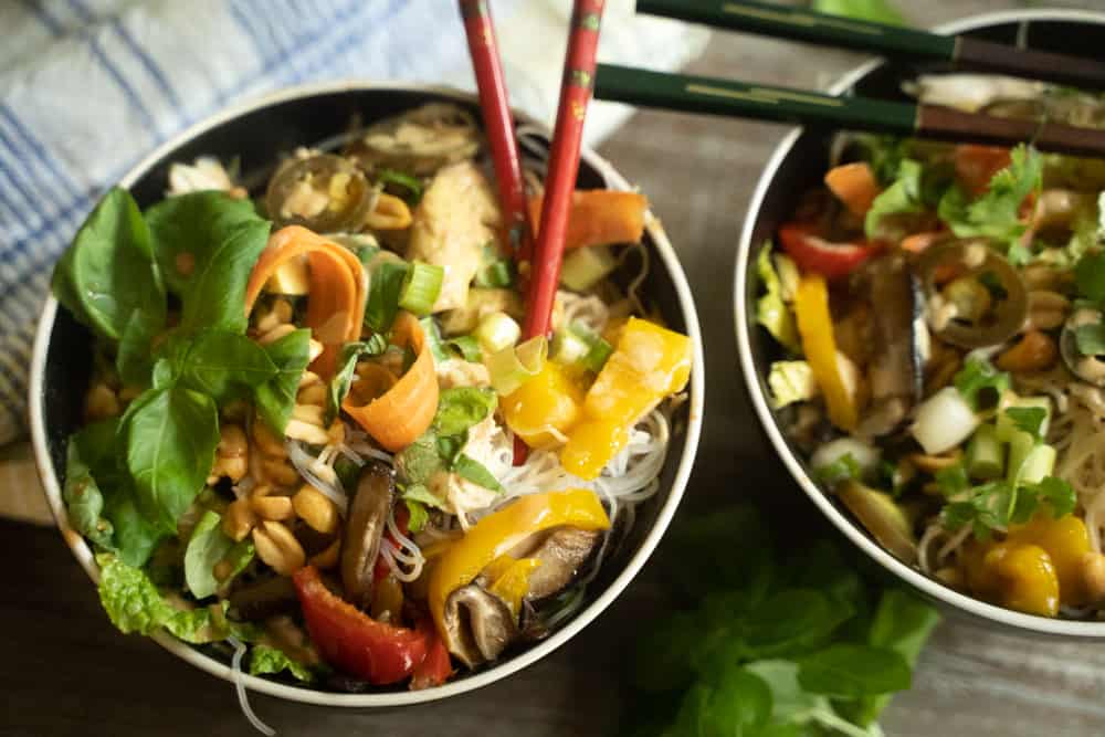 2 bowls filled with spring roll ingredients of shaved carrots, slices of yellow and red pepper, mushrooms, peanuts, green onions, rice noodles drizzled with peanut sauce with chopsticks resting against the bowls