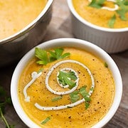 2 white bowls filled with creamy carrot soup swirled with sour cream and garnished with parsley sitting on a wood table next to a silver soup terrine