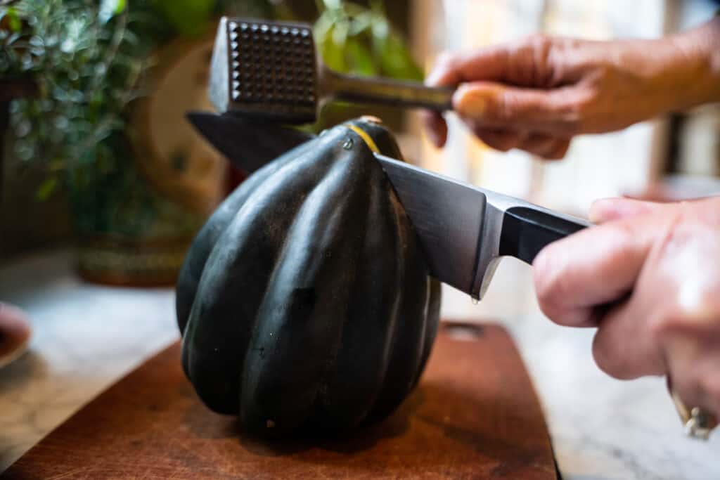 Acorn squash on a cutting board being sliced open with a chef knife tapped by a mallet.