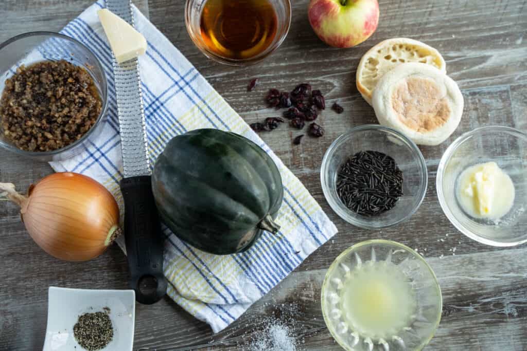 Ingredients for sausage stuffed acorn squash for two - acorn squash, english muffin, maple syrup, chicken broth, dried cranberries, onion, salt, pepper, apple, parmesan cheese on a table