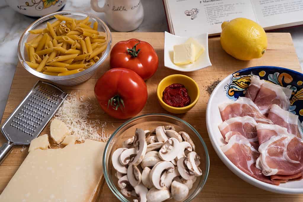 prosciutto pasta ingredients on a counter including penne, fresh tomatoes, mushrooms, butter, lemon, parmesean, prosciutto