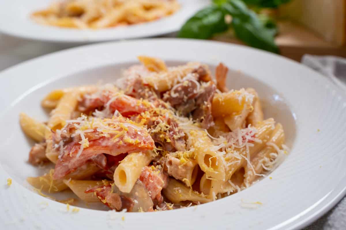 Proscuitto pasta with fresh tomatoes, mushrooms and cooked prosciutto garnished with parmesan cheese and lemon zest in a white bowl