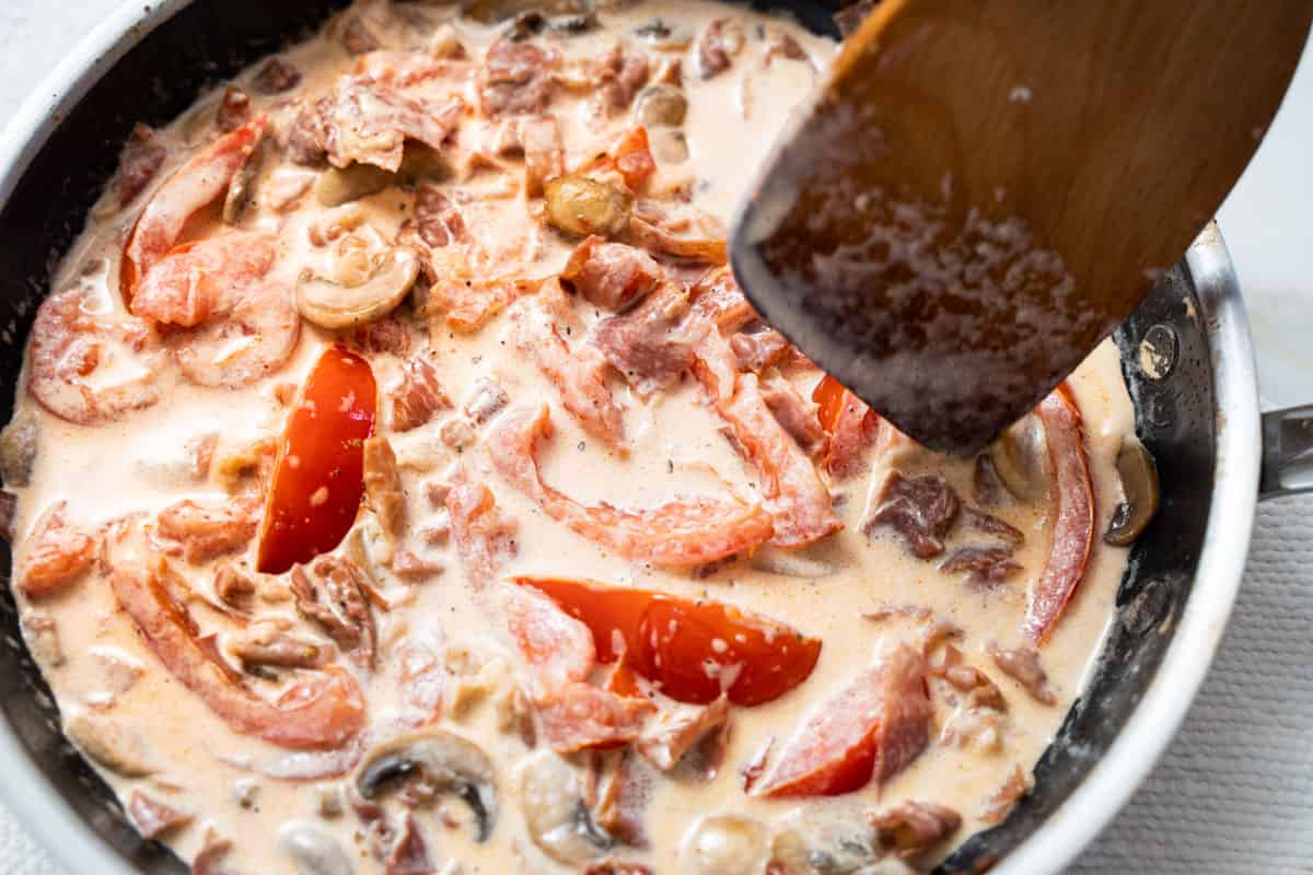 prosciutto pasta sauce in a skillet being stirred by a wooden spoon