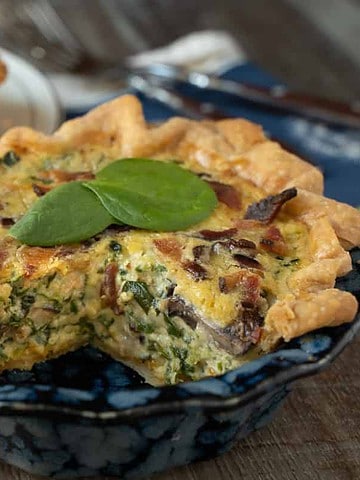 quiche for two made with egg, spinach, mushroom, cheese and bacon in a blue pie plate with one slice cut out