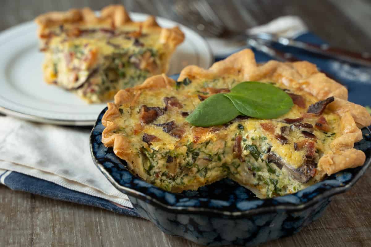 quiche for two made with egg, spinach, mushroom, cheese and bacon in a blue pie plate with one slice cut out