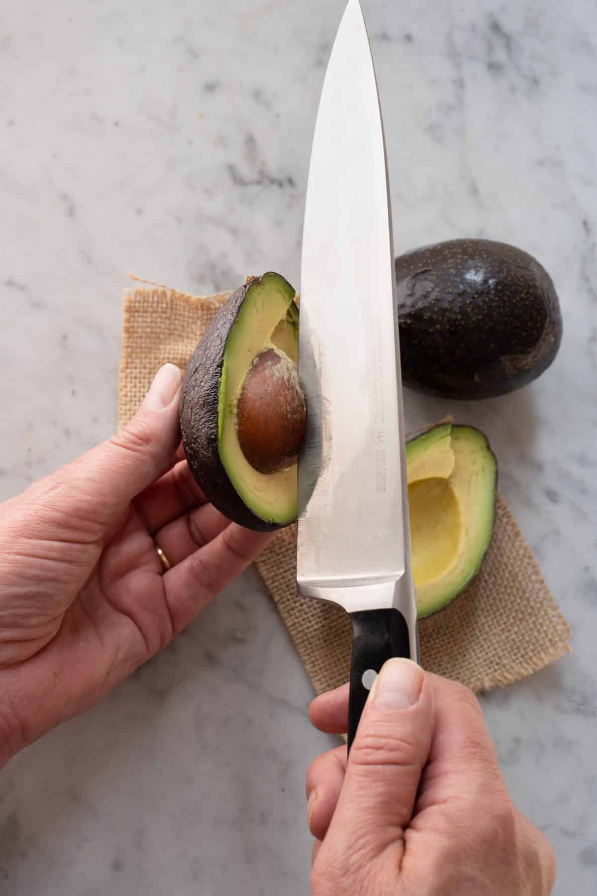 left hand holding avocado and the right hand holding a knife wedged into the pit of the avocado