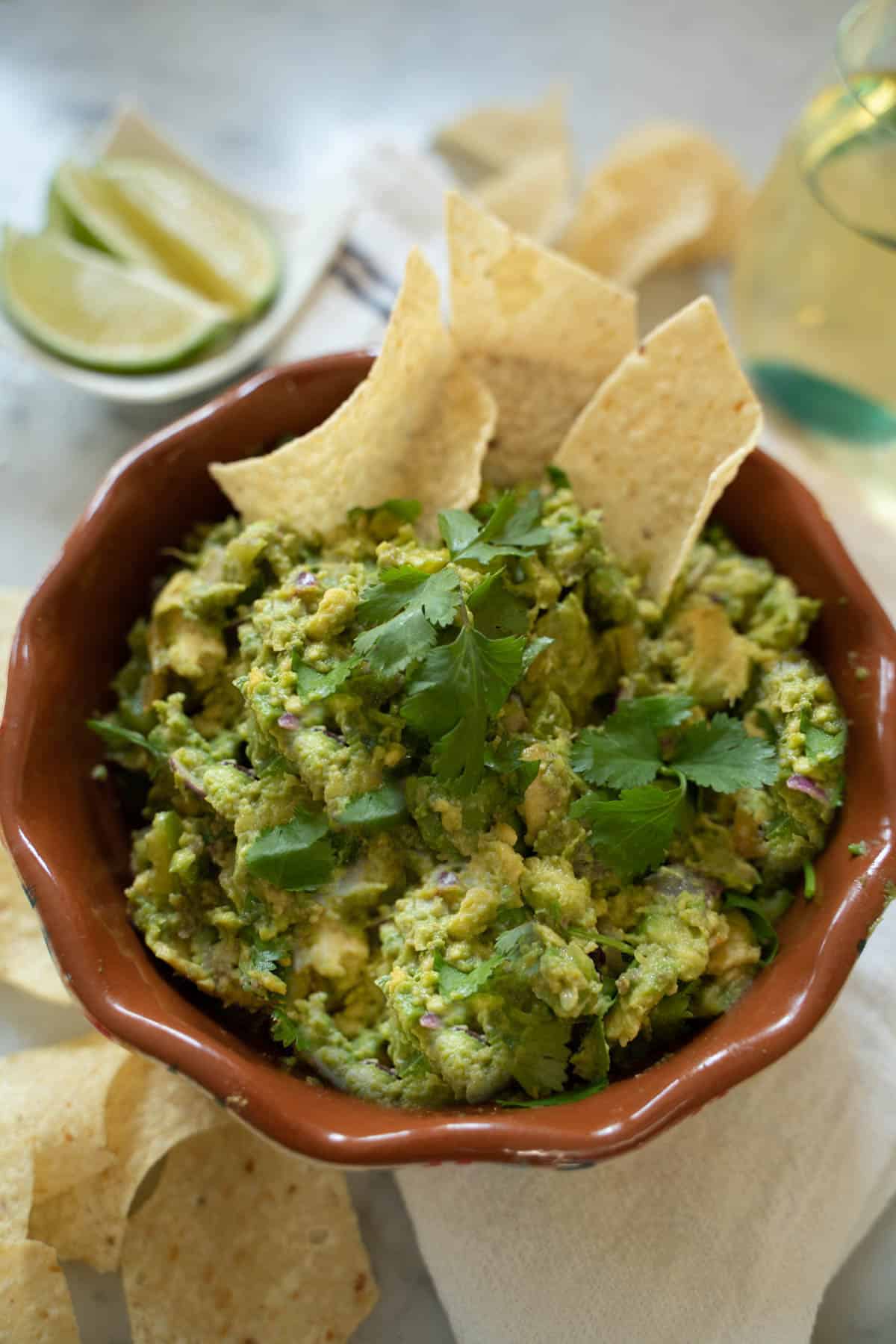 guacamole in a brown bowl surrounded by tortilla chips and a glass of white wine