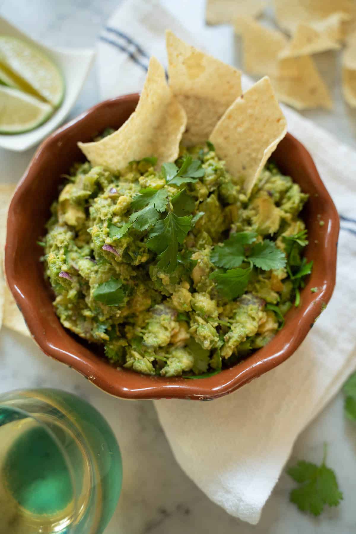Guacamole in a brown bowl surrounded by tortilla chips and a glass of white wine