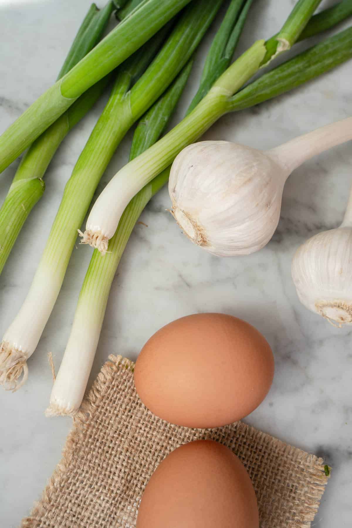 green onions, 2 eggs and heads of garlic on a marble countertop