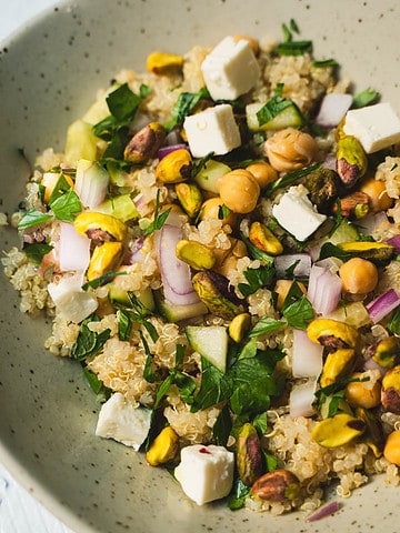 Jennifer Aniston Salad Recipe made of quinoa,cucumber, fresh herbs, pistachios, chickpeas and feta cheese all tossed in a simple lemon and olive oil dressing in a small bowl on a white table