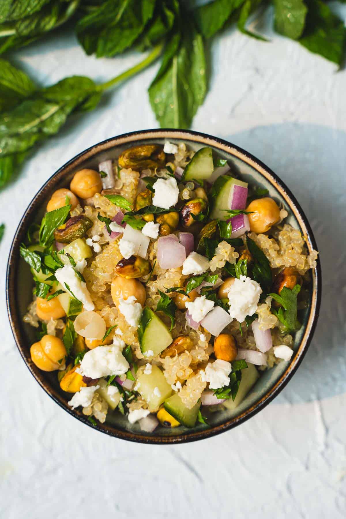 Jenniifer Aniston Salad Recipe made of quinoa,cucumber, fresh herbs, pistachios, chickpeas and feta cheese all tossed in a simple lemon and olive oil dressing in a small bowl on a white table