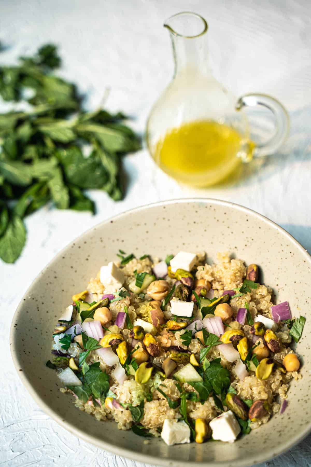 eniifer Aniston Salad Recipe made of quinoa,cucumber, fresh herbs, pistachios, chickpeas and feta cheese all tossed in a simple lemon and olive oil dressing in a green oval bowl on a white table with mint and olive oil bottle sitting next to the bowl
