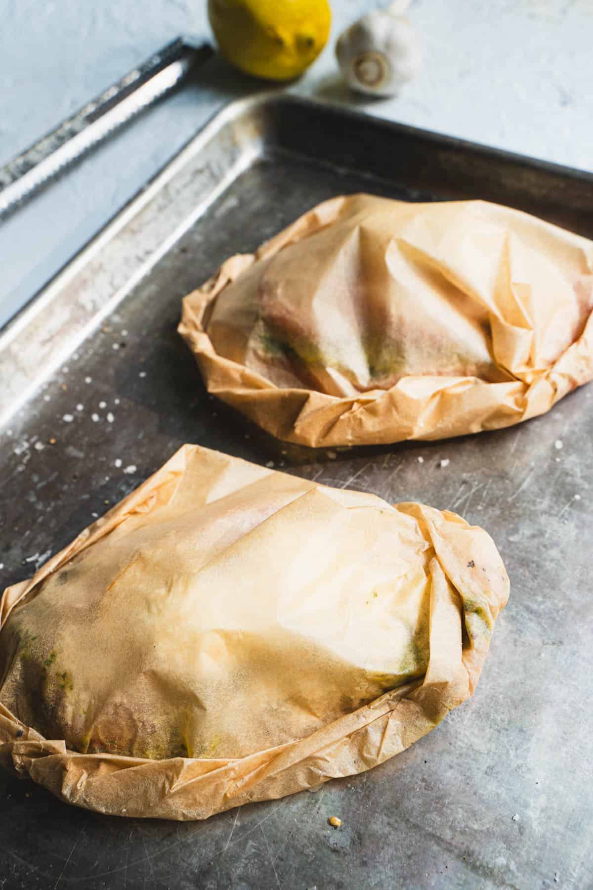 baked pesto chicken breasts and ingredients inside folded and crimped closed parchment bundles on a baking sheet.