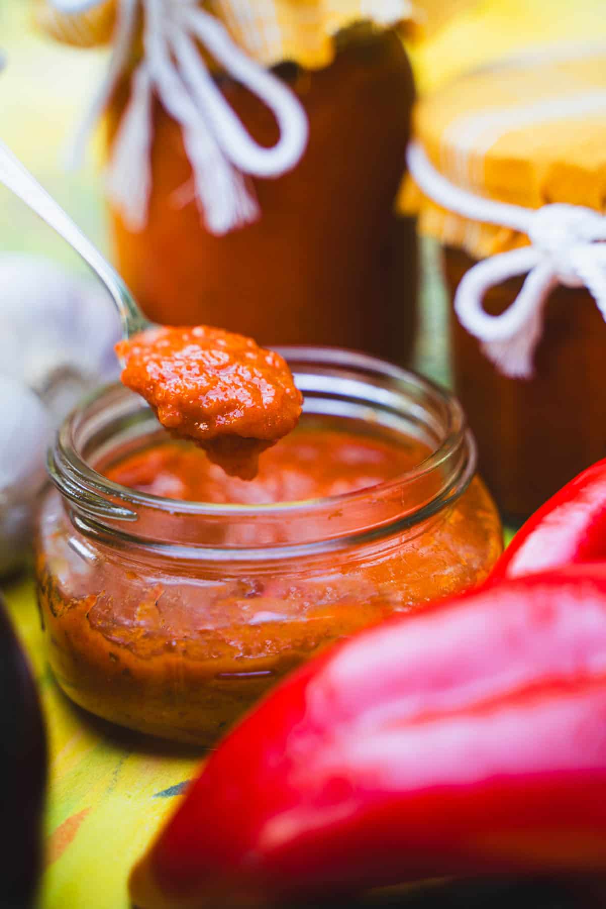 Ajvar dipping sauce in a wide mouth mason jar with a spoon surrounded by sealed mason jars and fresh vegetables