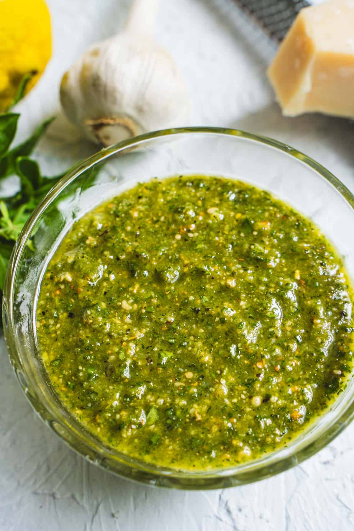 Classic fresh basil pesto in a glass bowl surrounded by ingredients to make the pesto on a white table