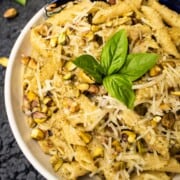 Pistachio Pesto Penne Pasta in a bowl sprinkled with parmesan and chopped pistachios with a basil garnish in a white bowl on a dark blue table