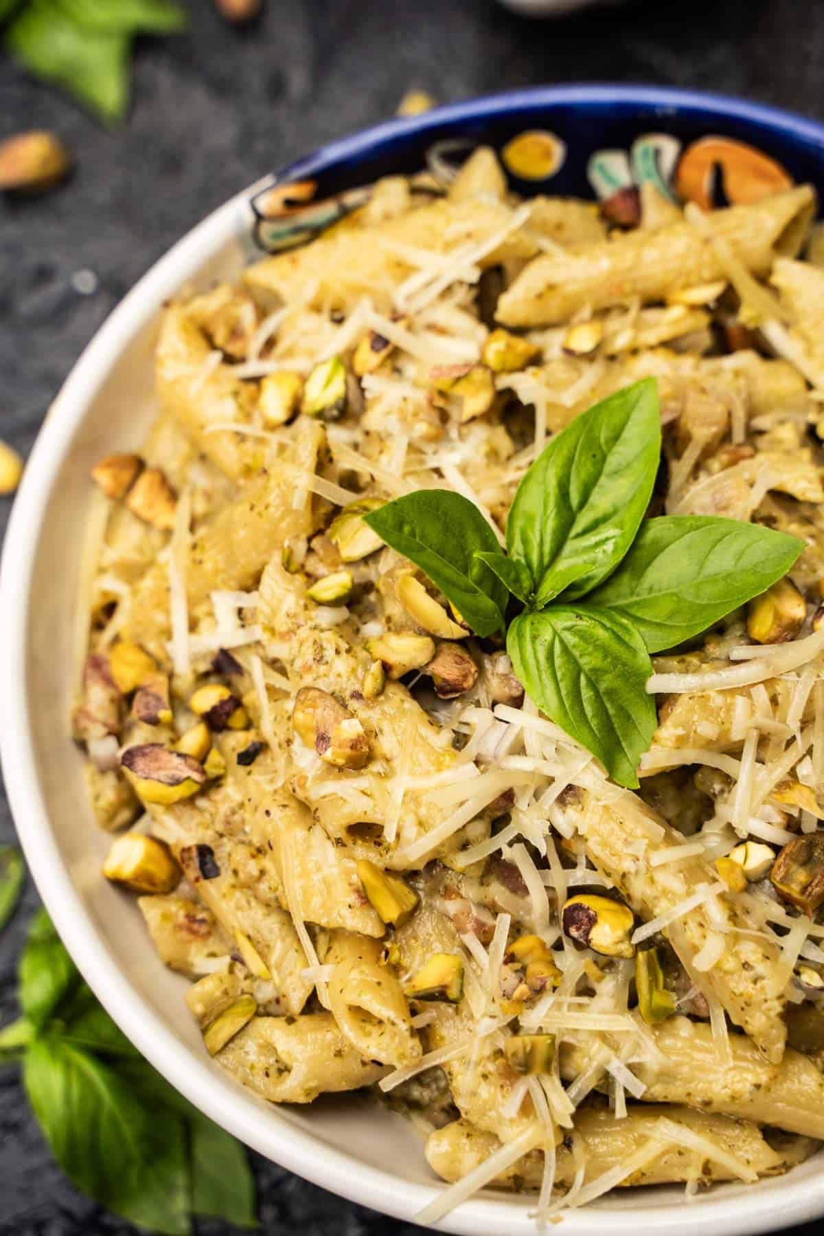 Pistachio Pesto Penne Pasta in a bowl sprinkled with parmesan and chopped pistachios with a basil garnish in a white bowl on a dark blue table