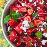 chunks of watermelon, sliced strawberries, slices of red onion, crumbled red onion and chopped mint in a bowl sitting on top of a sunflower tablecloth