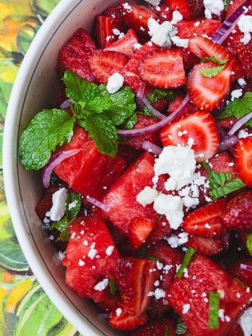 chunks of watermelon, sliced strawberries, slices of red onion, crumbled red onion and chopped mint in a bowl sitting on top of a sunflower tablecloth