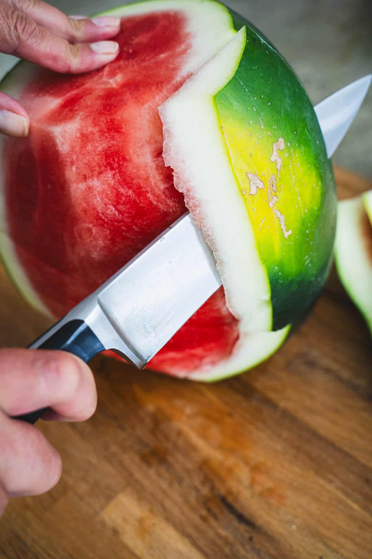 Watermelon on end with one hand holding the melon and one hand holding a chef's knife to remove rind