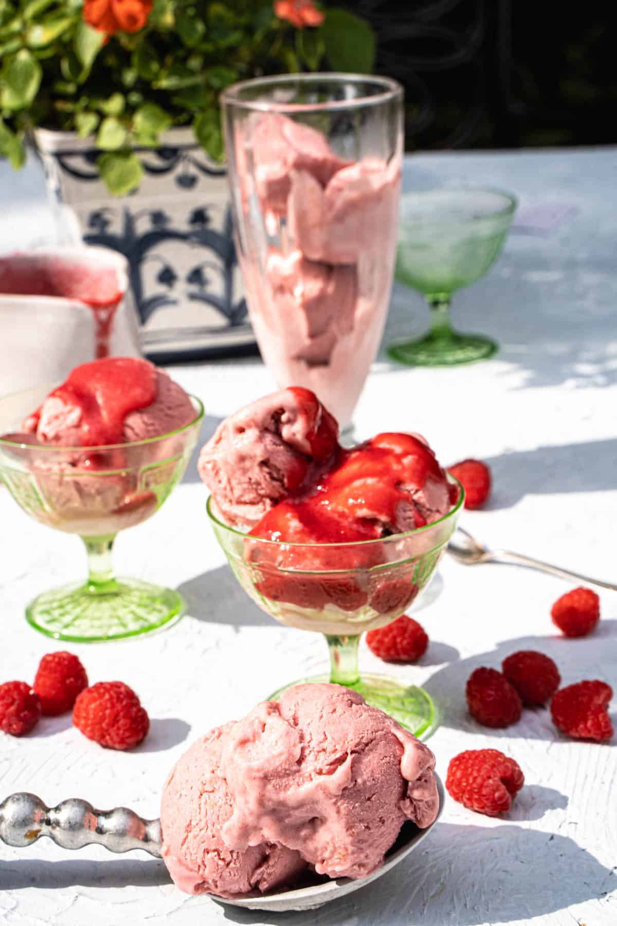 no churn Raspberry ice cream in green ice cream glass dessert cups and a clear tall ice cream glass and a scoop on a silver spoon sitting on a white table