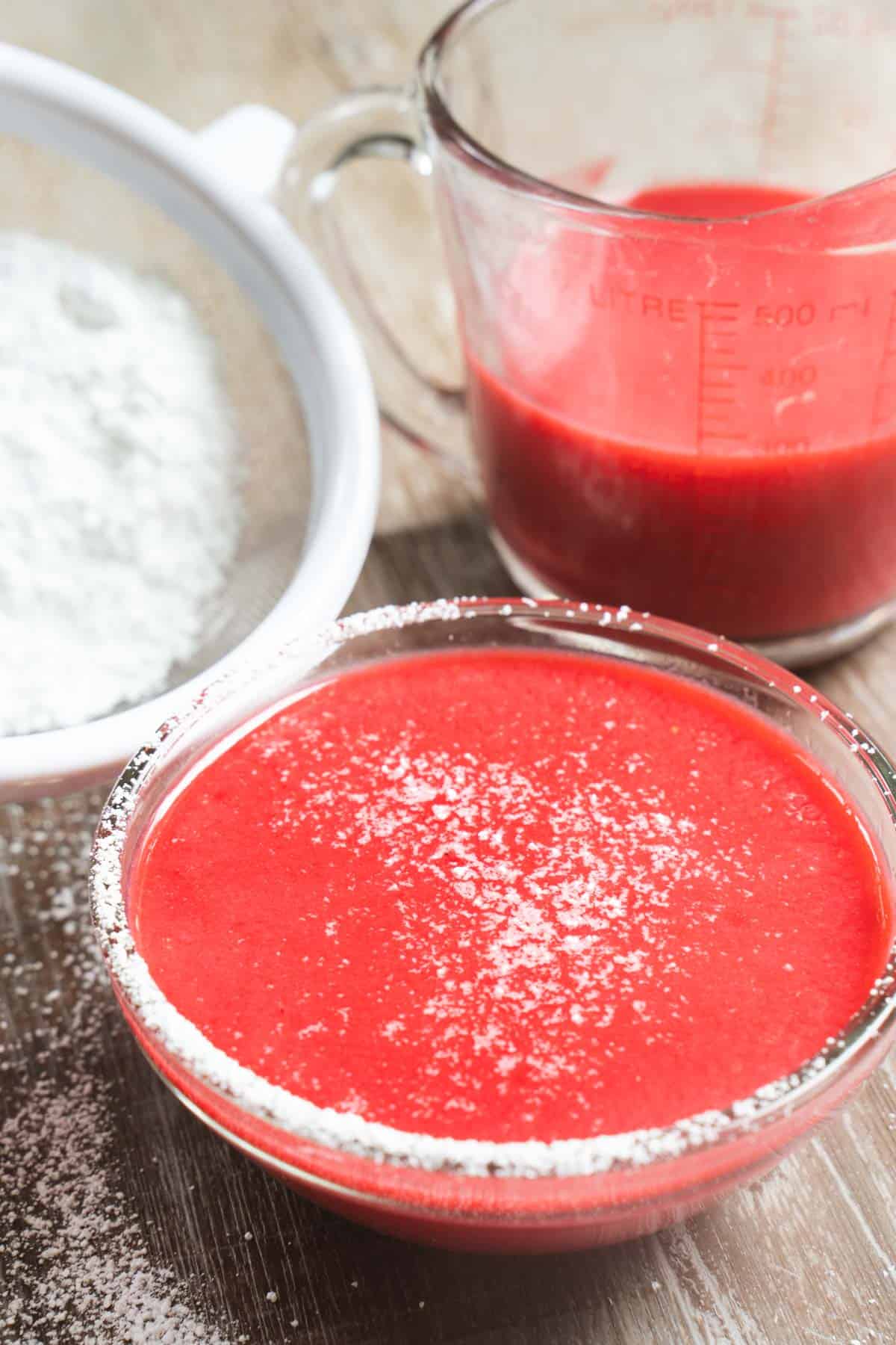 Raspberry puree in a bowl and in a 2 cup measuring cup along side a sieve filled with powdered sugar on a wooden table